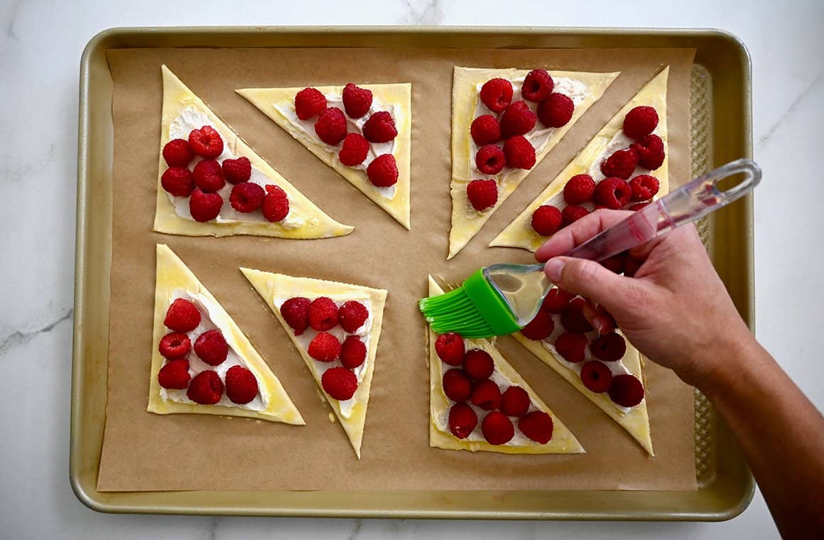 A hand holding a pastry brush applies an egg wash to triangle-shaped danish on a parchment paper-lined baking sheet