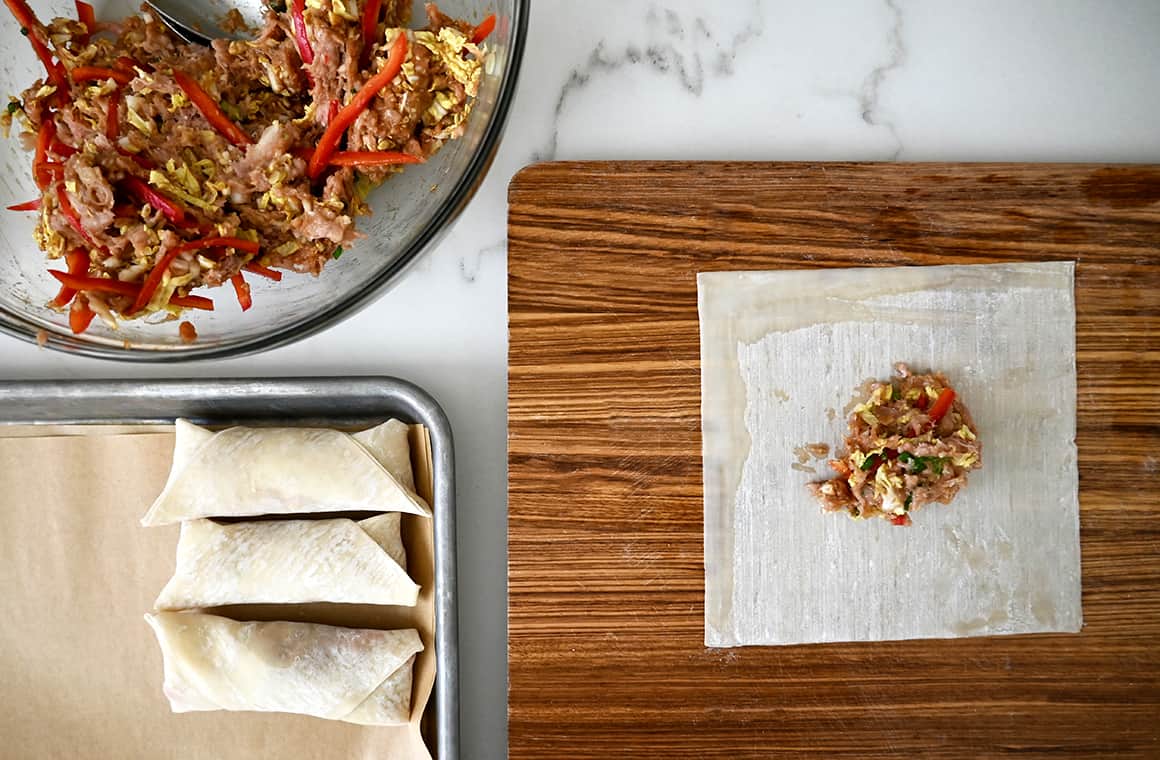 A top-down view of an egg roll wrapper with filling on a cutting board next to a baking pan with unbaked egg rolls and a clear bowl containing the filling