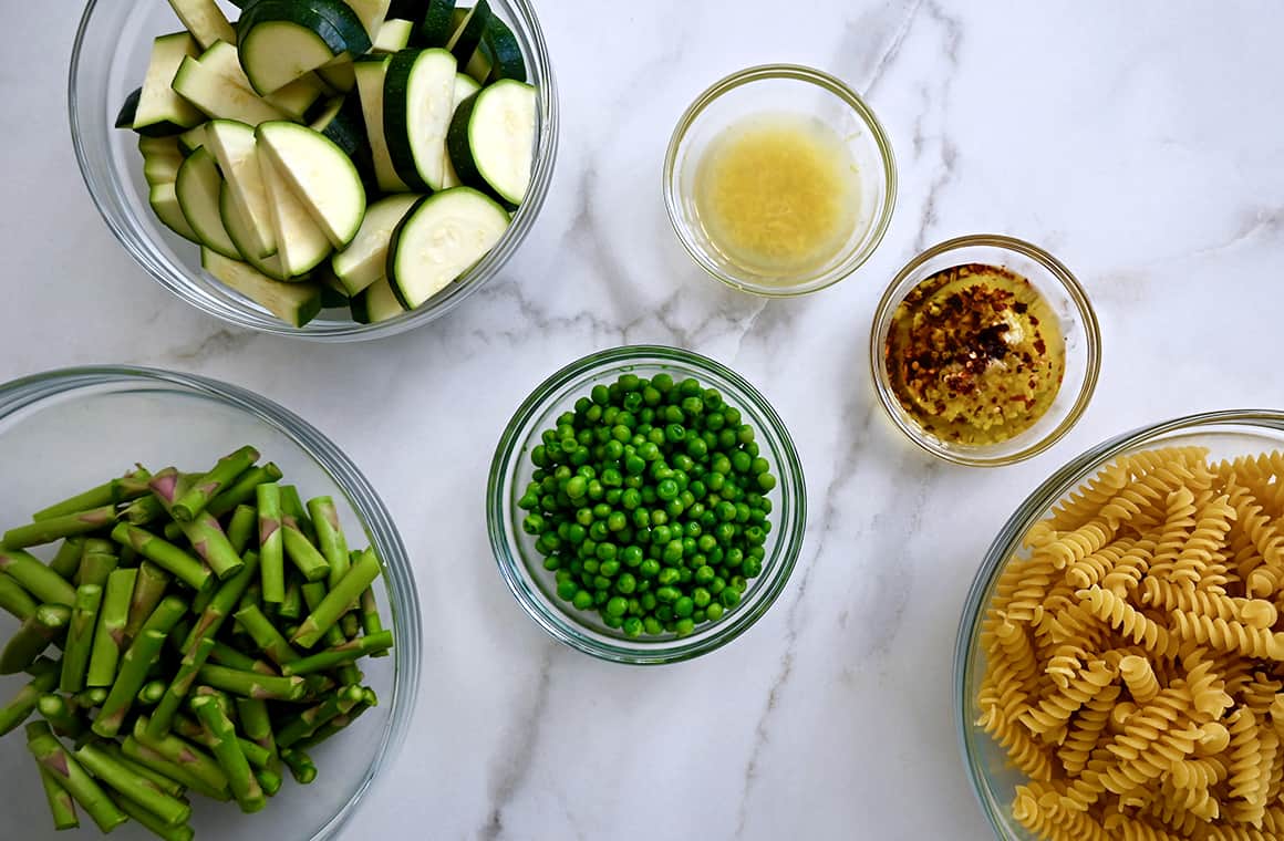 Various sizes of clear bowls containing asparagus pieces, sliced zucchini, peas, lemon juice, chili oil and uncooked rotini pasta