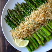 Roasted asparagus topped with cheesy breadcrumbs on a white plate with a lemon wedge.