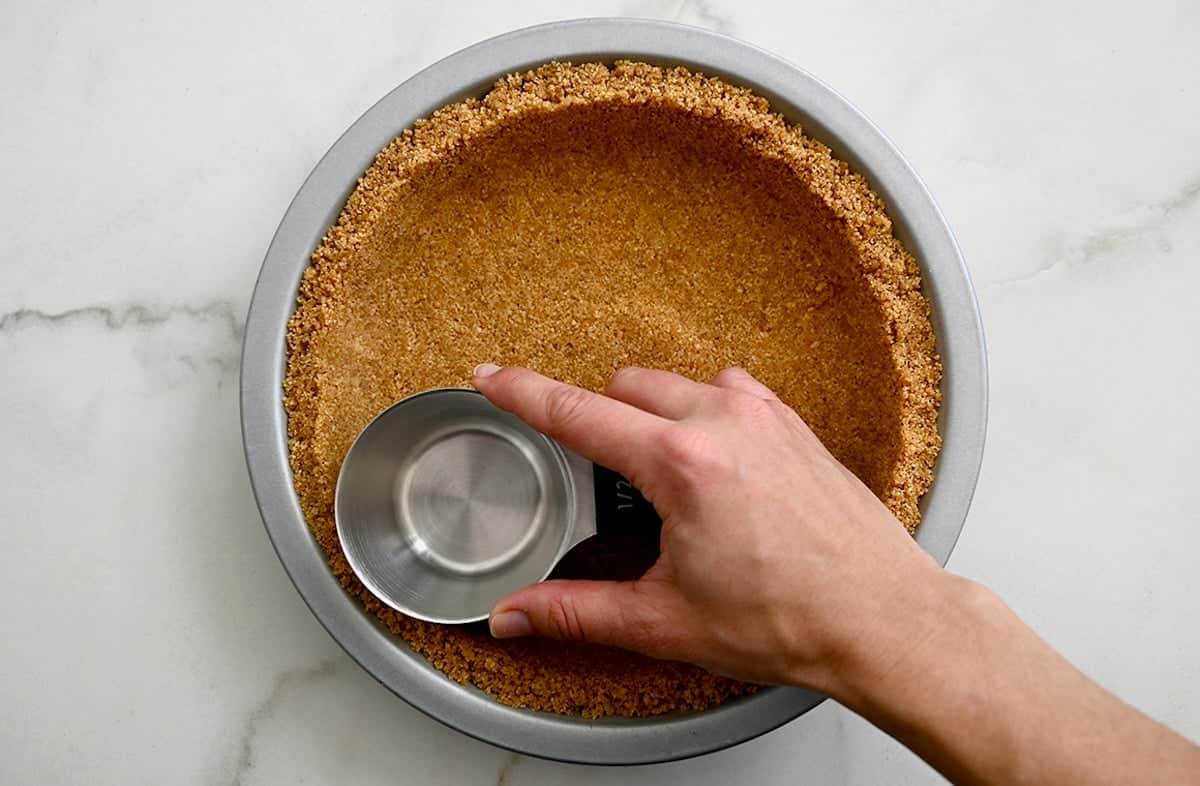 A hand holding a measuring cup presses graham cracker crumbs into a pie pan to form a graham cracker crust.