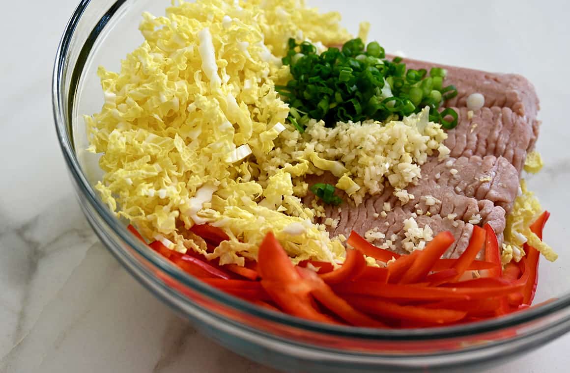 A clear bowl containing ground turkey, shredded cabbage, sliced red bell pepper, minced ginger and finely chopped scallions