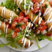 A top-down view of air fryer cheese taquitos topped with pico de gallo and guacamole on a white plate atop a bed of lettuce.