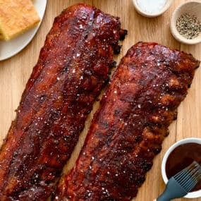A top-down view of Oven-Baked Baby Back Ribs brushed with barbecue sauce next to a plate with cornbread and a bowl containing sliced pickles