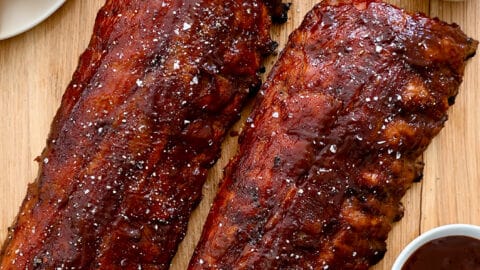 A top-down view of Oven-Baked Baby Back Ribs brushed with barbecue sauce next to a plate with cornbread and a bowl containing sliced pickles