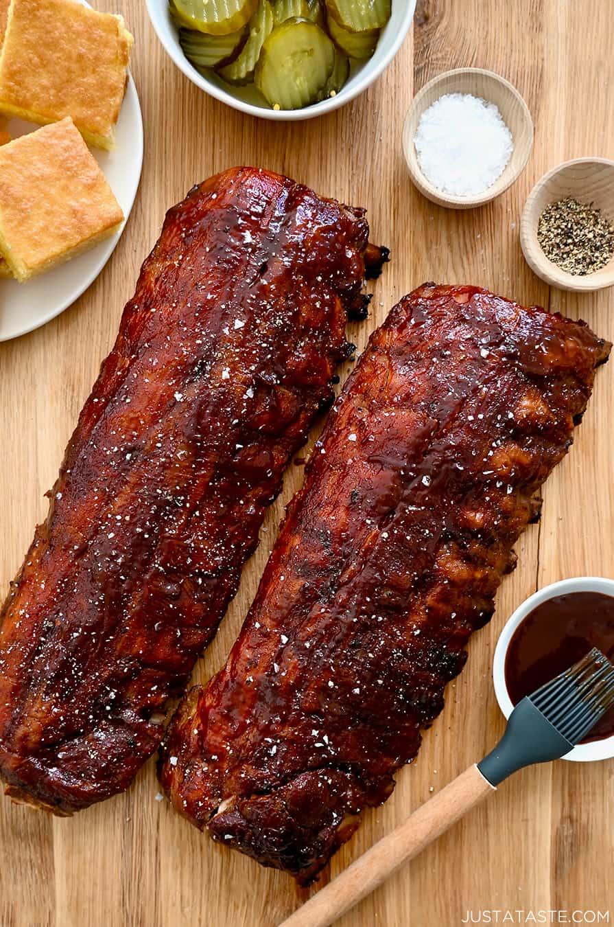 A top-down view of Oven-Baked Baby Back Ribs brushed with barbecue sauce next to a plate with cornbread and a bowl containing sliced pickles