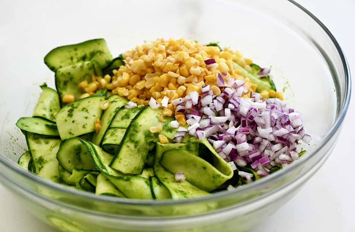 A clear bowl containing zucchini ribbons, chopped red onion and corn topped with a basil vinaigrette