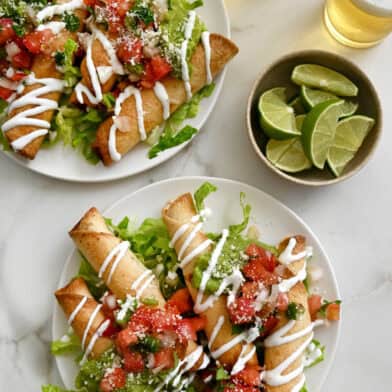 Two plates containing crispy chicken taquitos atop a bed of shredded lettuce and topped with pico de gallo and a drizzle of sour cream.