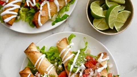 Two plates containing crispy chicken taquitos atop a bed of shredded lettuce and topped with pico de gallo and a drizzle of sour cream.
