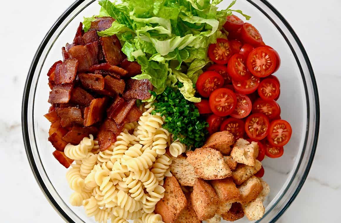 A top-down view of a clear bowl containing chopped bacon, lettuce, halved cherry tomatoes, croutons and rotini