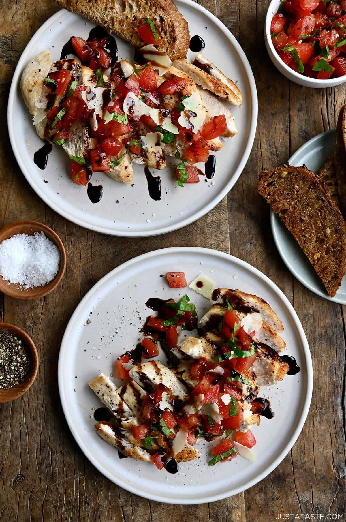 Two plates containing seared chicken breasts topped with bruschetta, shaved Parmesan and a drizzle of balsamic syrup.