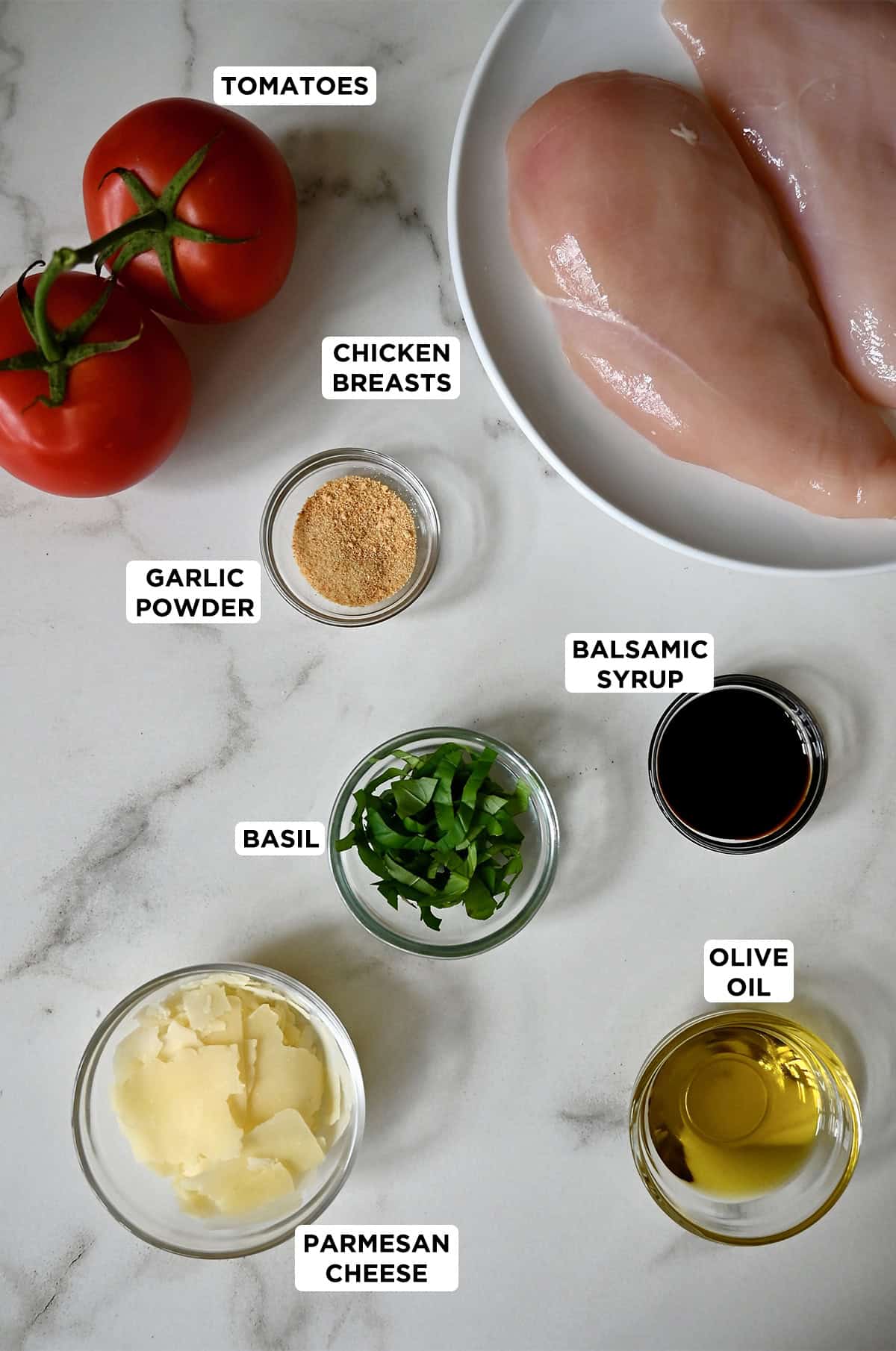 Boneless, skinless chicken breasts on a plate next to bruschetta ingredients, including tomatoes, garlic powder, fresh basil and shaved parmesan cheese. Two small bowls containing olive oil and balsamic syrup are nearby.