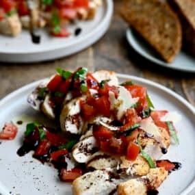 Easy bruschetta chicken topped with shaved Parmesan cheese and a drizzle of balsamic syrup on a white dinner plate. A plate piled high with crusty bread slices is nearby.