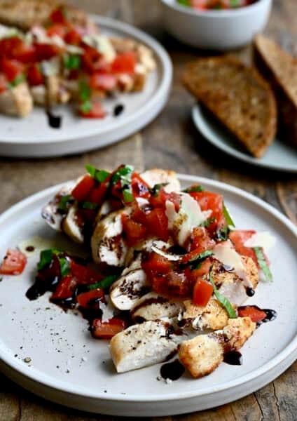 Easy bruschetta chicken topped with shaved Parmesan cheese and a drizzle of balsamic syrup on a white dinner plate. A plate piled high with crusty bread slices is nearby.