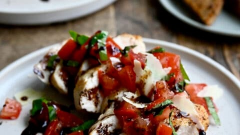Easy bruschetta chicken topped with shaved Parmesan cheese and a drizzle of balsamic syrup on a white dinner plate. A plate piled high with crusty bread slices is nearby.
