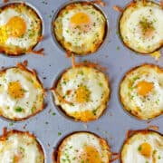 A top-down view of a muffin pan containing cheesy hash brown cups with baked eggs garnished with salt, pepper and chives.