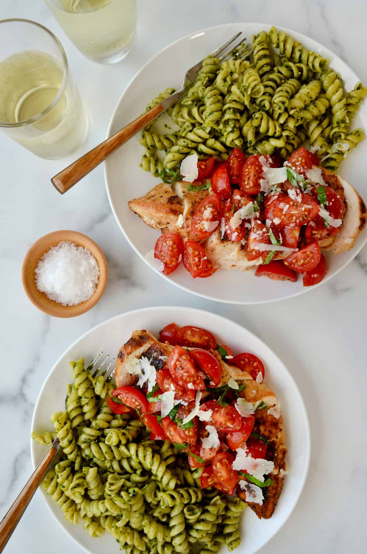 Two plates containing chicken breasts with a bruschetta topping and pesto pasta. 