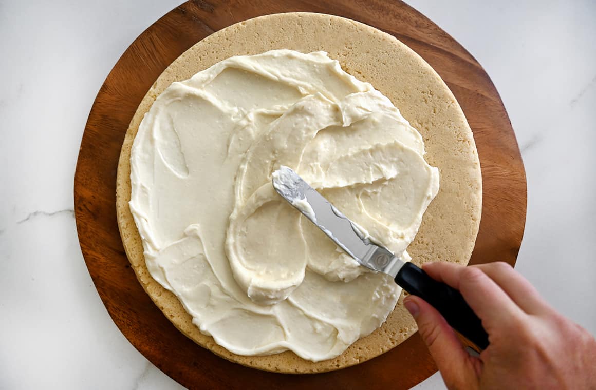 A hand holding an offset spatula spreads vanilla cream cheese frosting atop a sweet crust