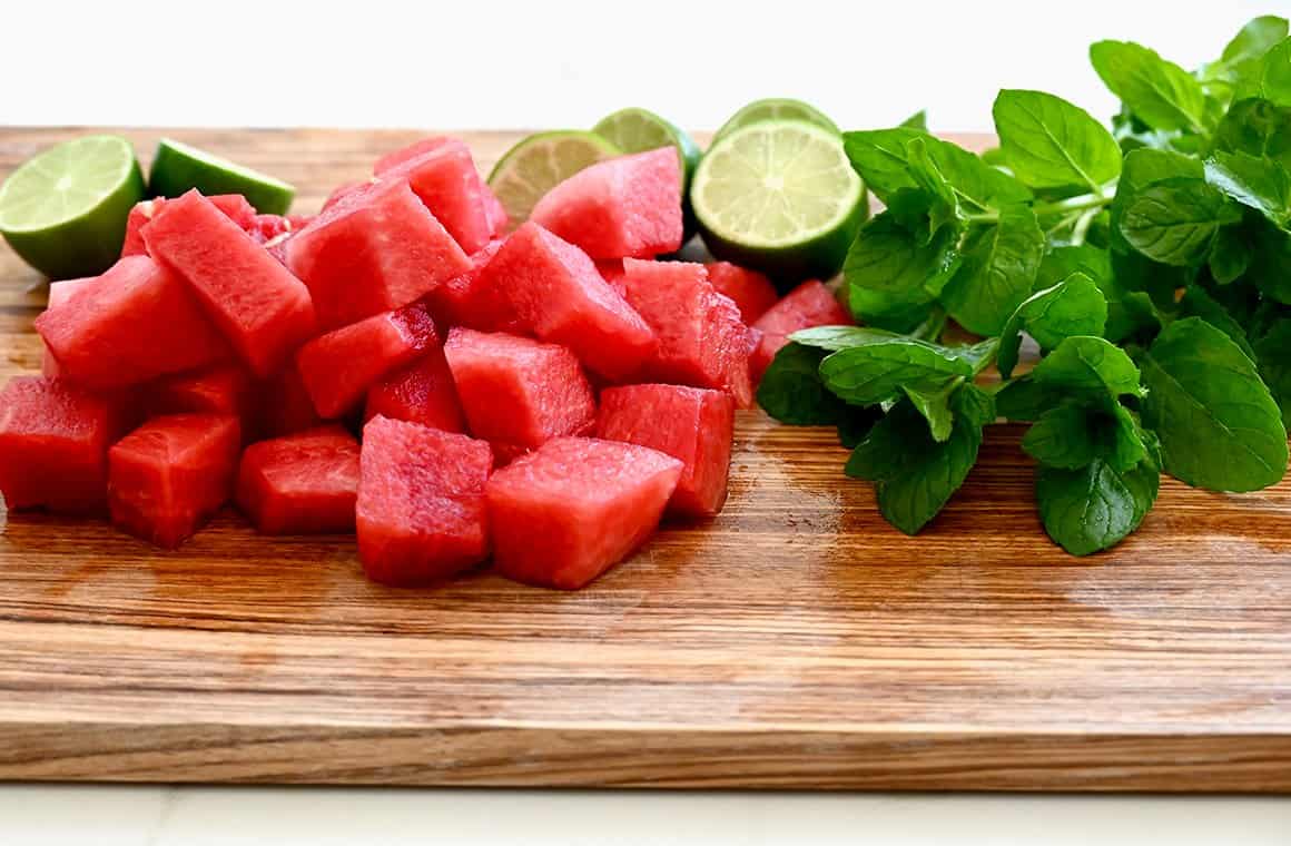 Mint leaves, cubed watermelon and halved limes on a wood cutting board