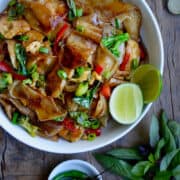 A top-down view of drunken noodles with chicken and fresh Thai basil in a white bowl next to birds eye chilies in a small bowl and a sprig of basil leaves.