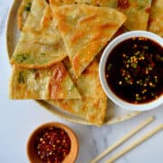 A top-down view of a small bowl filled with crushed red pepper flakes and chopsticks next to a plate topped with flaky scallion pancakes and a bowl containing soy dipping sauce.