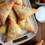 Strawberry Rhubarb Turnovers piled on a white serving plate next to a small bowl filled with sanding sugar and a bowl containing fresh strawberries.