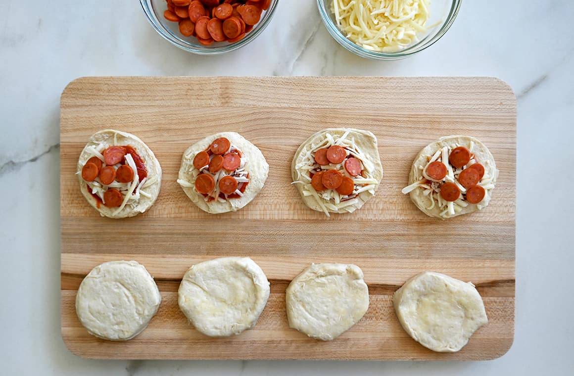 A wood cutting board containing biscuit dough rounds filled with marinara and pizza toppings