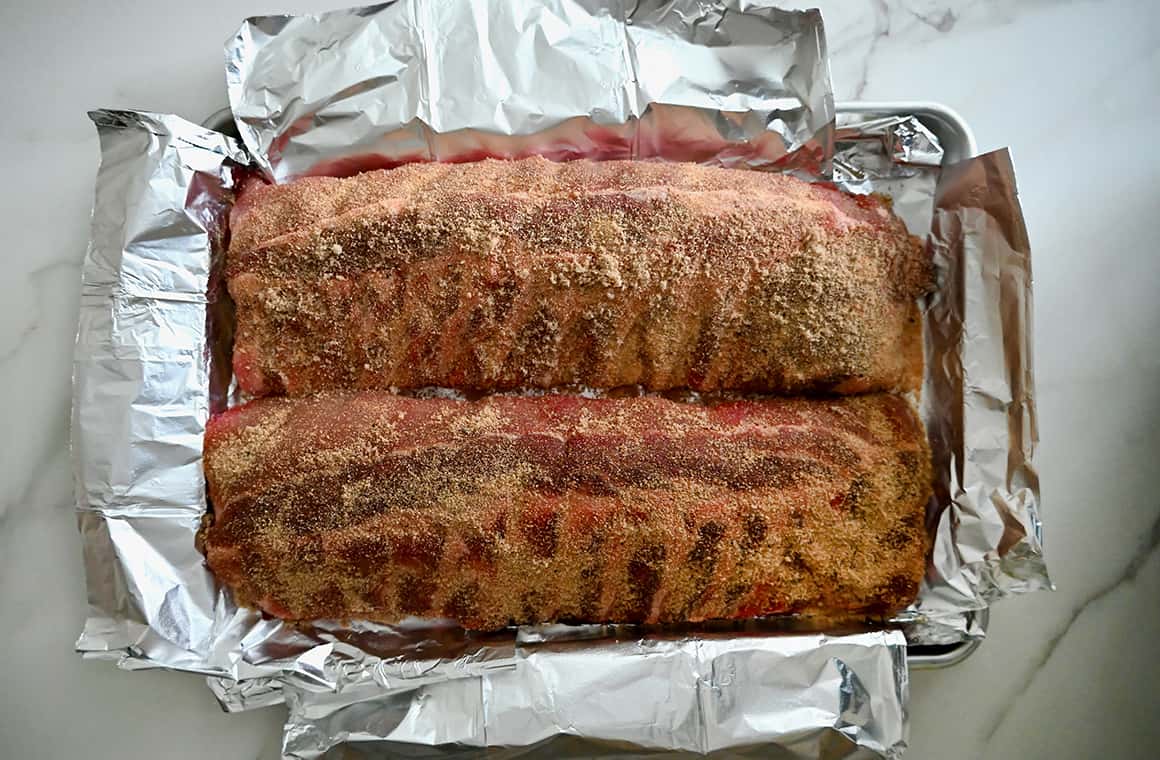 Unbaked rib slabs with a dry spice rub atop a foil-lined baking sheet