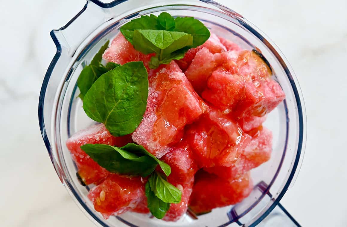 A top-down view of frozen melon, honey and mint leaves in a blender