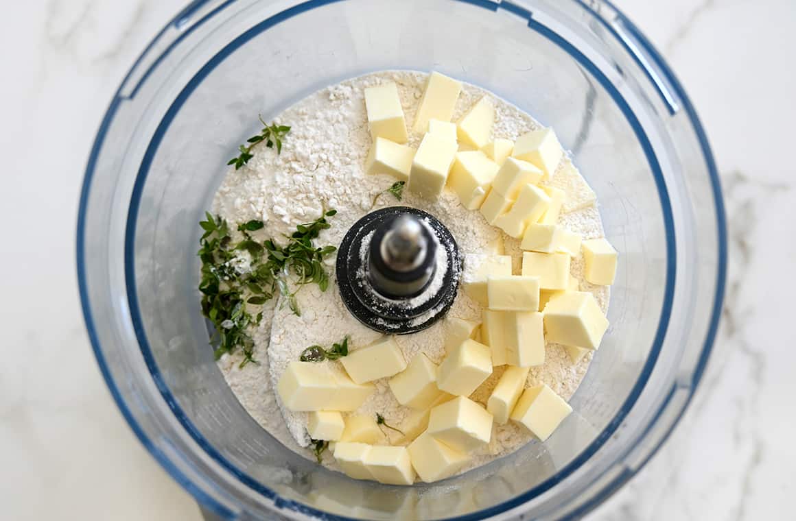A top-down view of a food processor bowl containing cubed butter, flour, sugar and fresh thyme