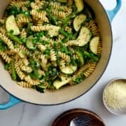 A top-down view of a stockpot containing Garlicky Pasta Primavera with zucchini, asparagus and peas next to small bowls containing fresh herbs and Parmesan cheese.