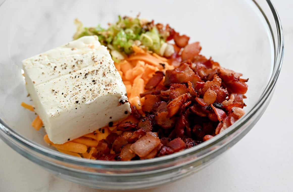 A clear bowl containing cream cheese, sliced scallions, shredded cheddar cheese and bacon pieces