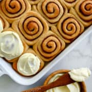 A top-down view of golden-brown cinnamon rolls in a white baking dish next to a bowl filled with creamy cream cheese frosting.