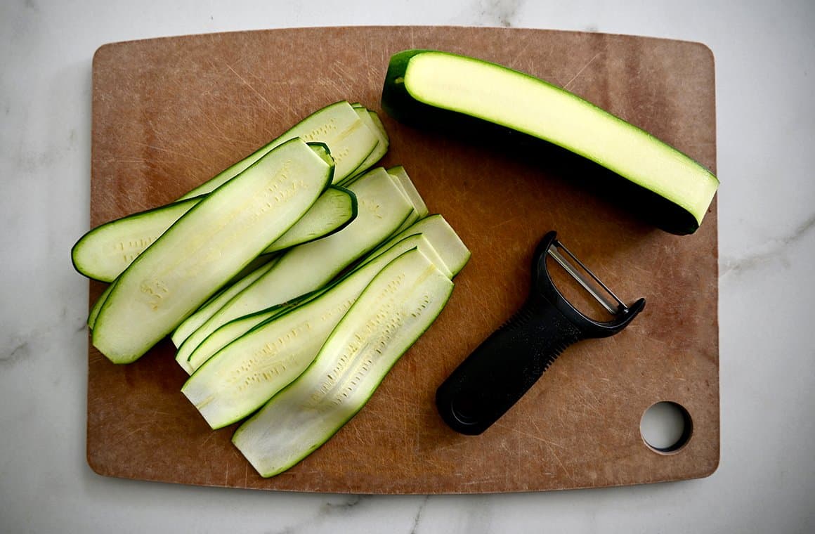 A top-down view of zucchini ribbons and a vegetable peeler atop a wood cutting board