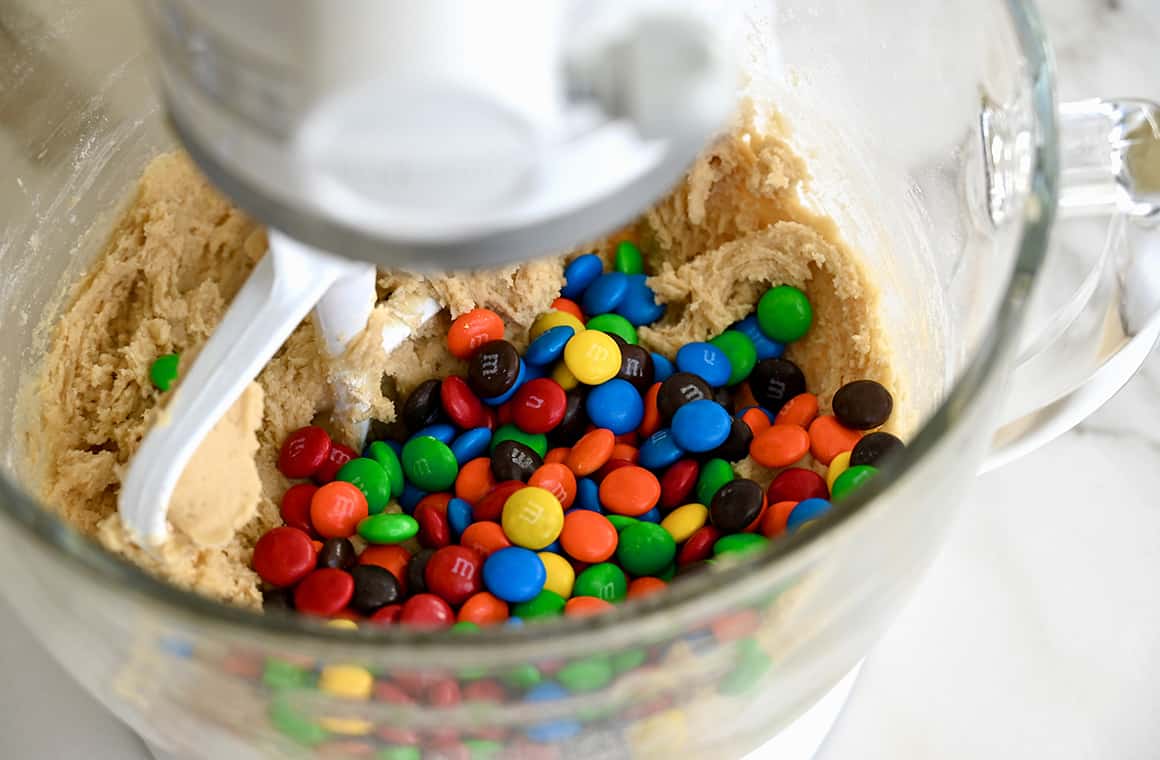 A stand mixer bowl containing dough with rainbow-colored candy-coated chocolates