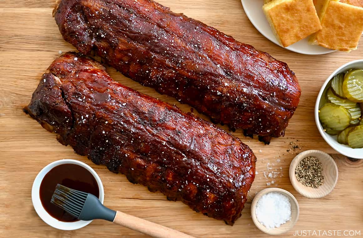 The best Oven-Baked Baby Back Ribs next to a small bowl containing barbecue sauce and a plate piled with cornbread