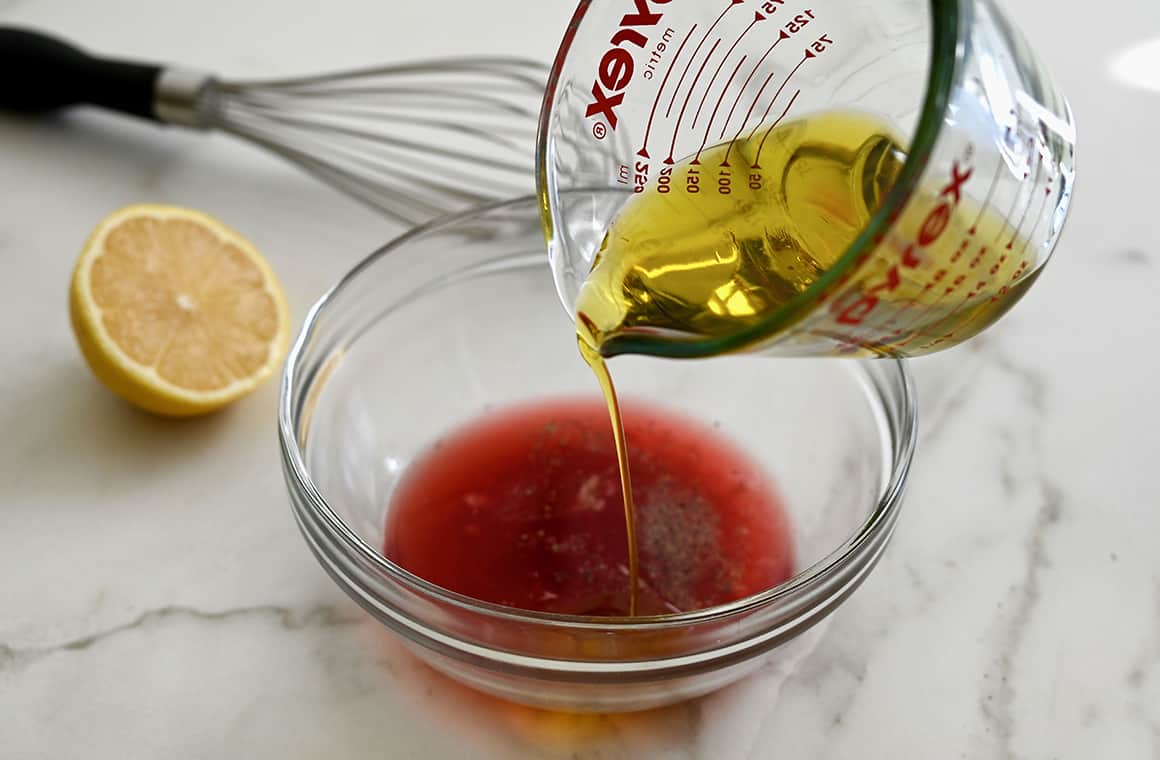 Olive oil in a glass measuring cup being poured into a clear bowl containing red wine vinegar and fresh lemon juice