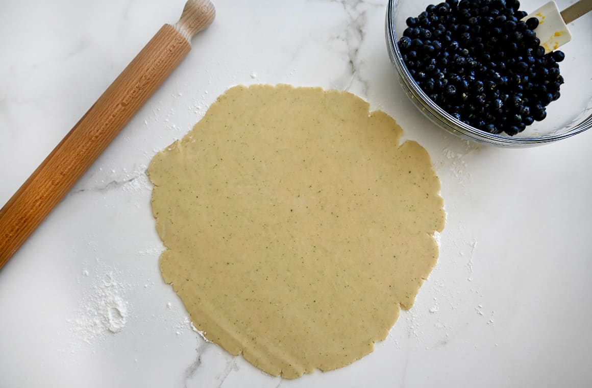 A top-down view of rolled-out pie dough next to a rolling pin and a clear bowl containing berries