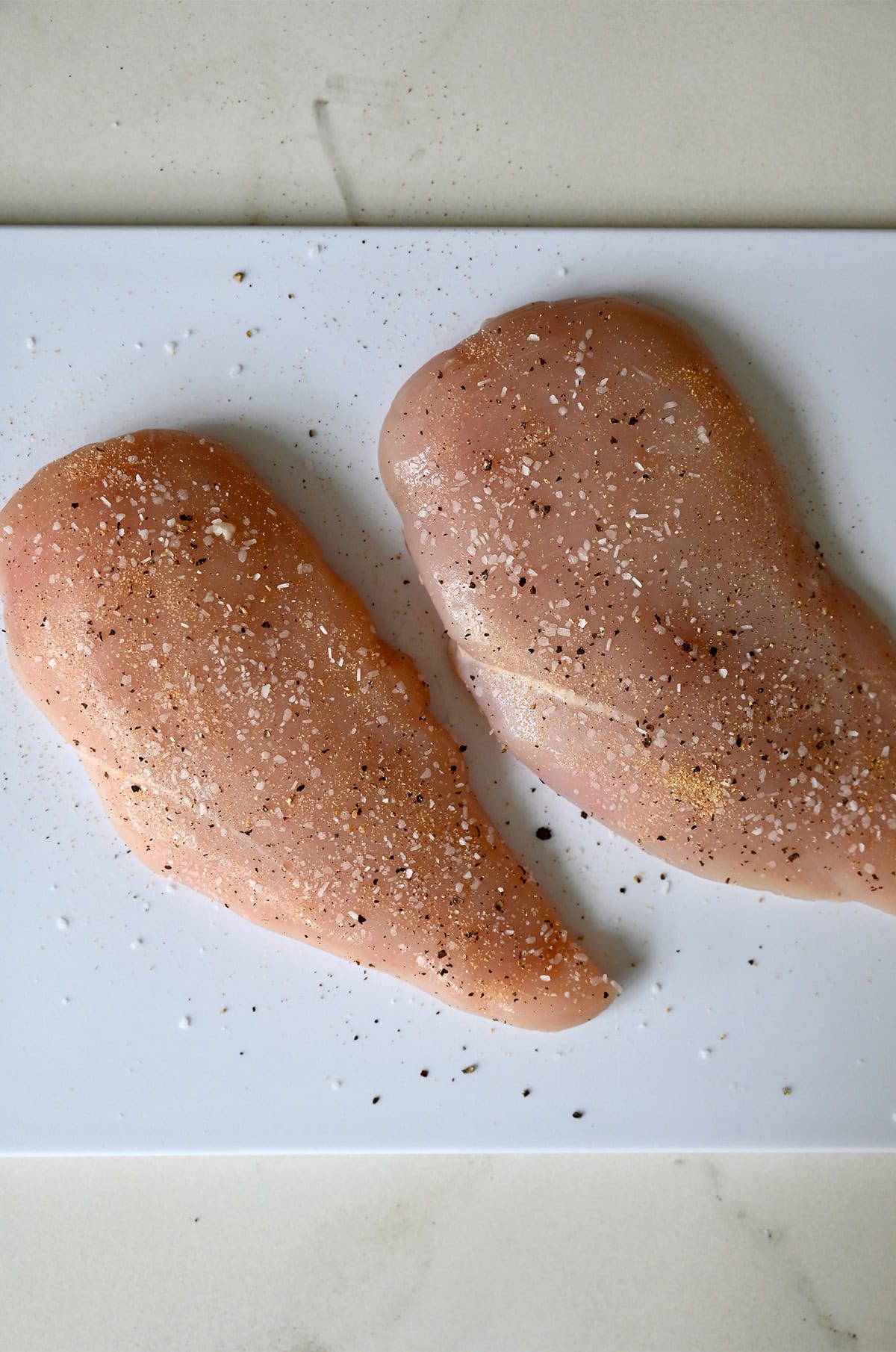 Two boneless, skinless chicken breasts on a cutting board seasoned with salt, pepper and garlic powder.