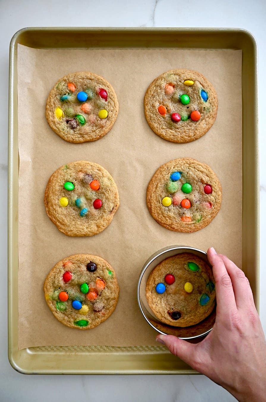 A top-down view of a hand holding a circular cookie cutter over a hot cookie to form into the perfect circle