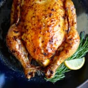 A top-down view of a whole roast chicken with fresh rosemary sprigs and a lemon wedge in a cast-iron skillet.