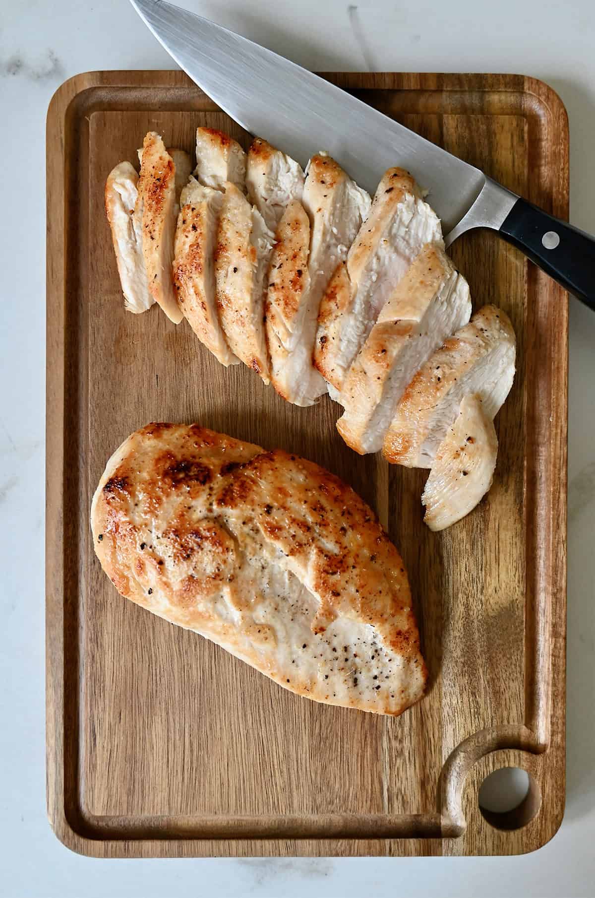 A seared and sliced chicken breast on a cutting board with another seared chicken breast and a sharp knife.