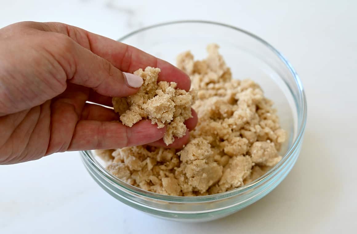 A hand holds streusel topping over a small bowl containing streusel