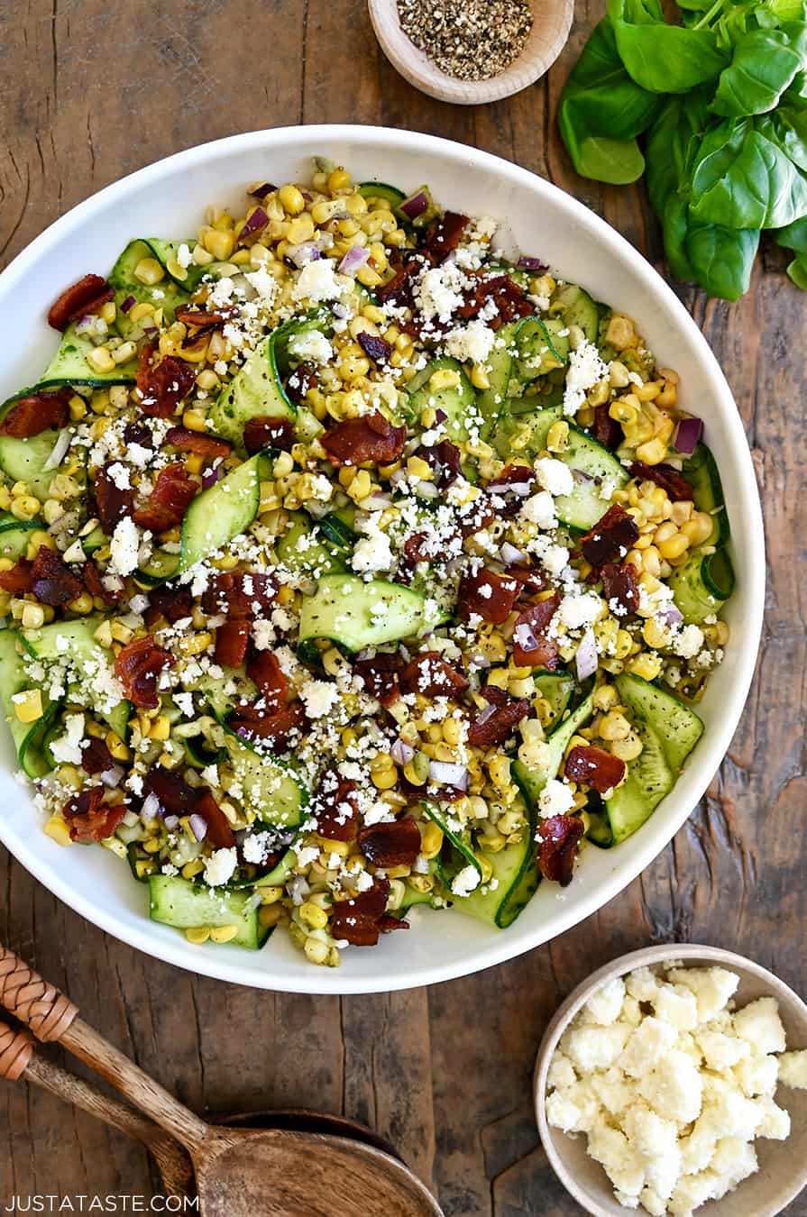 A top-down view of Corn and Zucchini Salad with Basil Vinaigrette in a large white serving bowl next to a small bowl filled with cotija cheese