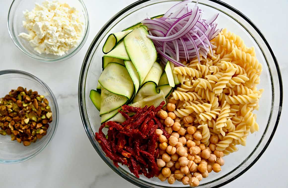 A large clear bowl containing cooked rotini noodles, chickpeas, sun-dried tomatoes, zucchini ribbons and sliced red onions next to two small bowls containing pistachios and feta cheese