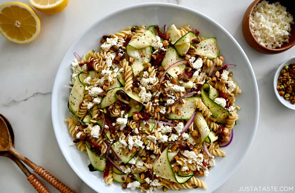 A top-down view of homemade zucchini pasta salad with feta cheese, sun-dried tomatoes, chickpeas and pistachios in a large white bowl