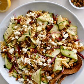 A top-down view of Zucchini Pasta Salad with feta cheese, chickpeas, sun-dried tomatoes and pistachios in a large serving bowl with wooden spoons