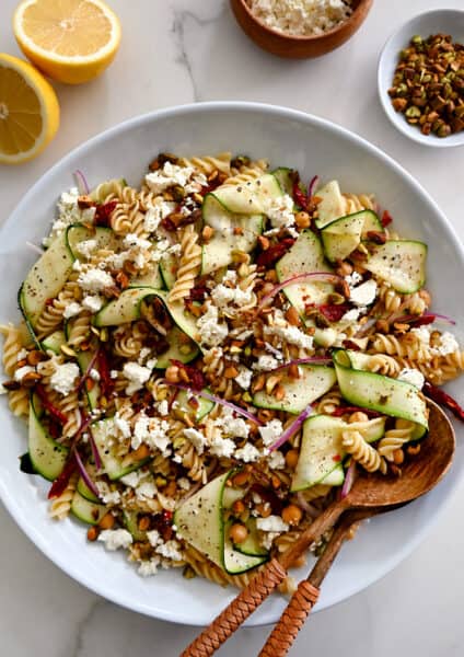 A top-down view of Zucchini Pasta Salad with feta cheese, chickpeas, sun-dried tomatoes and pistachios in a large serving bowl with wooden spoons