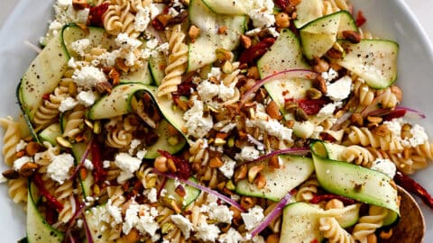 A top-down view of Zucchini Pasta Salad with feta cheese, chickpeas, sun-dried tomatoes and pistachios in a large serving bowl with wooden spoons