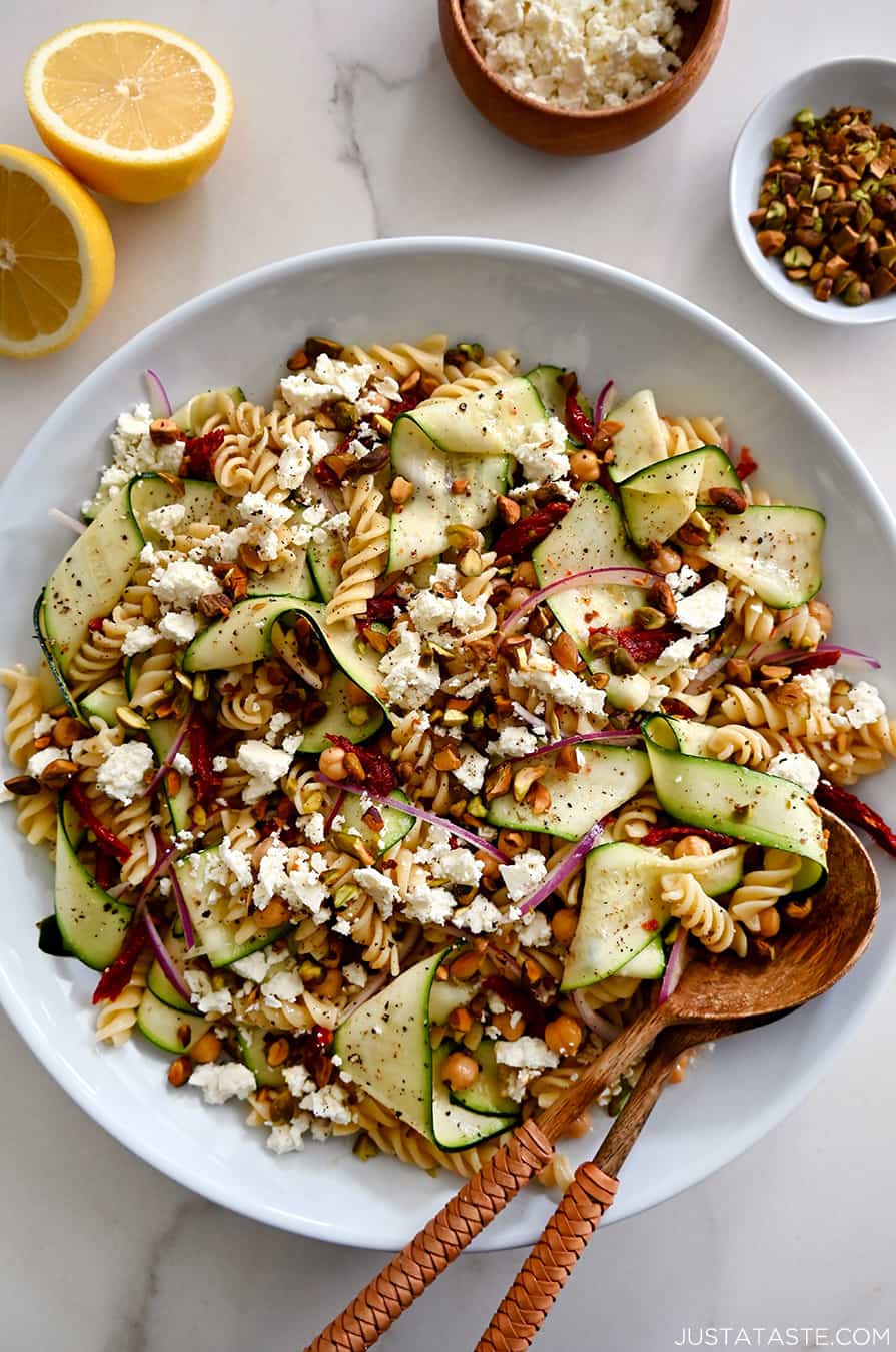 A top-down view of Zucchini Pasta Salad with feta cheese, chickpeas, sun-dried tomatoes and pistachios in a large serving bowl with wooden spoons